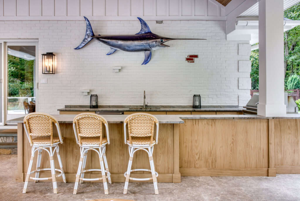 Wide view of an outdoor kitchen with U‑shaped stone counters, stainless steel grill, natural wood cabinetry, and a forested backdrop.