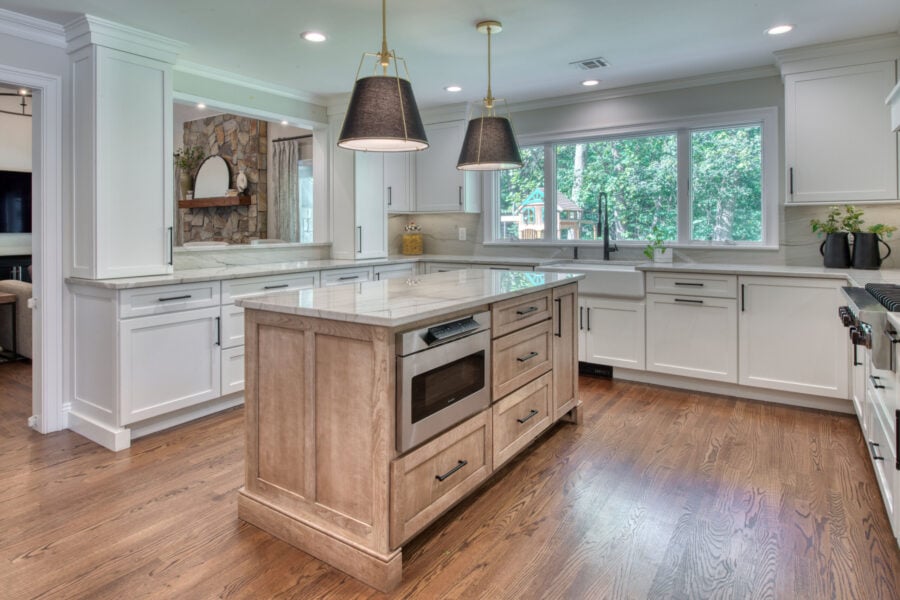 Kitchen island view showcasing a wood-tone island with built-in microwave, surrounded by white cabinetry and large windows.