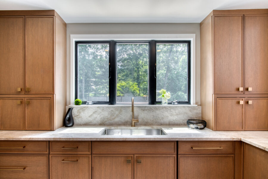 Kitchen sink area with a wide window, rift‑cut oak cabinets, stone backsplash, and minimalist countertop décor.