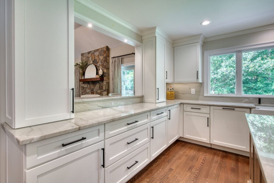 White shaker kitchen with marble-look counters, wide upper pass-through opening, and a view into a living space with a stone fireplace.