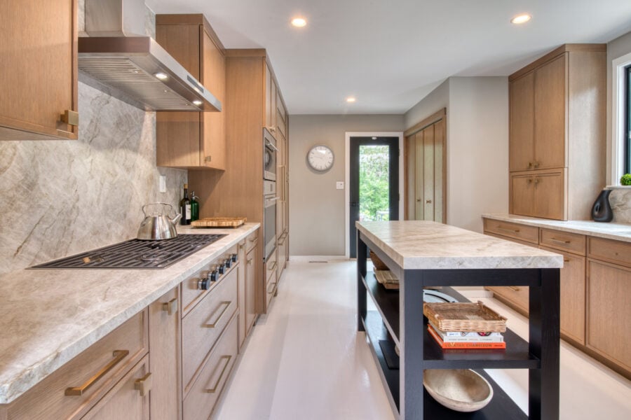 Wide view of a modern kitchen with rift‑cut oak cabinetry, stone countertops, stainless cooktop, and a contrasting dark island.