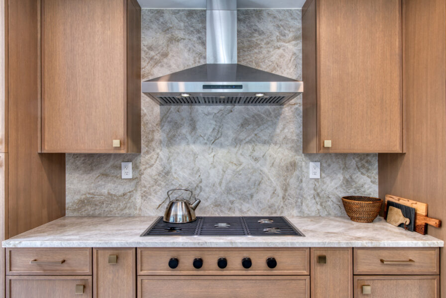 Cooktop area with stone backsplash, stainless hood, and rift‑cut oak cabinetry in a modern kitchen.