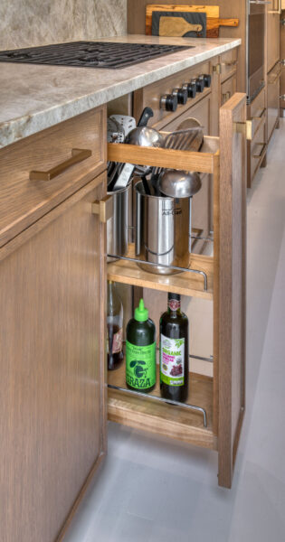 Pull‑out base cabinet organizer holding cooking utensils, olive oil, and vinegar bottles in a rift‑cut oak kitchen.